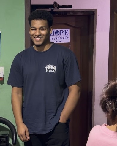 Shawn smiling in the same classroom, a HOPE worldwide Nepal sign on the door behind him